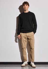 Pull en tricot noir, chinos beiges et baskets multicolores. Texture lisse, encolure ronde et logo de marque visible sur le pull.