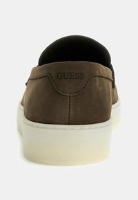 Zapato de ante marrón tipo slip-on con punta redondeada, suela de goma blanca y logo embossé en el talón. Textura suave y diseño minimalista.