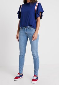 Top azul marino con mangas con volantes y detalles a rayas rojas y blancas, combinado con jeans ajustados de color azul claro y zapatillas rojas y azules.