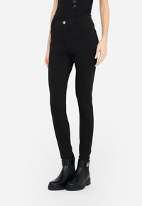 Pantalons ajustés noirs avec une texture lisse, dotés d'un bouton doré et d'un design minimaliste, portés avec des bottines noires.