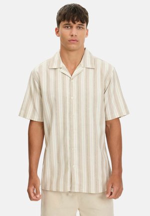 Joven de pie vistiendo una camisa beige de manga corta con rayas verticales y pantalones beige sobre un fondo liso.