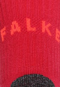 Röd strumpa med texturerad väv, med orange "FALKE"-bokstäver. Svarta häl- och tåsektioner med en slät yta.