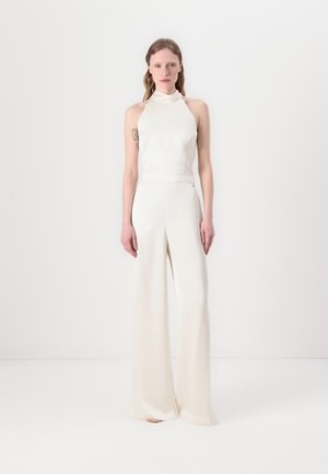 Weißer Jumpsuit mit Halterneck und weitem Hosenbein, aus glattem Stoff, mit minimaler Detailgestaltung, und einem Akzent auf der linken Hüfte.