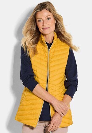 Blonde Frau mit lockigem Haar, die eine gelbe Steppweste über einem navyblauen Langarmhemd trägt und mit verschränkten Armen vor einem weißen Hintergrund steht.