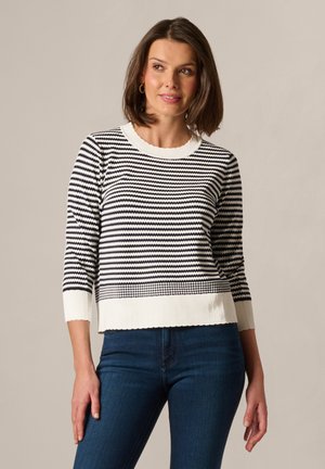 Frau mit schulterlangem braunem Haar, die einen schwarz-weiß gestreiften Pullover mit 3/4-Ärmeln und dunkelblaue Jeans trägt, schaut leicht nach rechts.