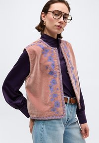 Γυναίκα με μοβ βελούδινο γιλέκο κεντημένο με ροζ λουλούδια πάνω από σκούρο μοβ μπλούζα, ανοιχτό μπλε τζιν και γυαλιά, ποζάρει με το χέρι στη μέση.