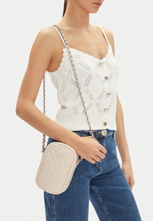 Frau trägt weißes ärmelloses Oberteil mit Knöpfen und blaue Jeans, hält beige gesteppte Umhängetasche mit Kettengurt.