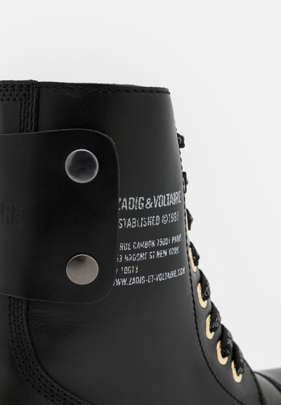 Black leather ankle boot with gold eyelets and black laces. Features a side strap with a snap button and embossed branding on the heel.