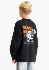 Zwart oversized langarm T-shirt met een kleurrijke cartoonbeerafbeelding en tekst op de achterkant, gemaakt van katoenen stof.