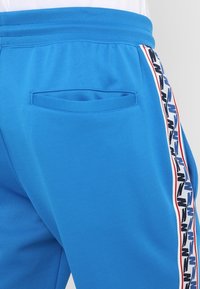 Modré sportovní šortky s boční kapsou a kontrastním proužkem s vzorem podél strany. Látka má hladkou strukturu.