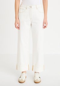 Mujer con vaqueros blancos de talle alto y pierna ancha con dobladillos, y zapatillas beige-blancas, de pie frente a un fondo claro y liso.