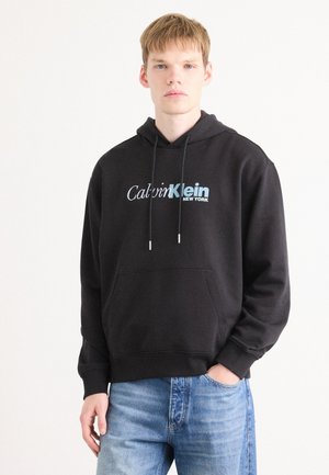 Black hoodie made of cotton, featuring a front pocket, adjustable drawstrings, and a blue logo that reads "Calvin Klein NEW YORK."