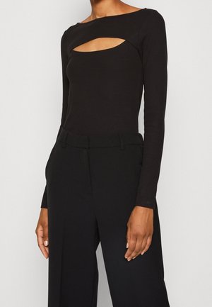 Femme portant un haut noir ajusté à manches longues avec une découpe à l'avant et un pantalon noir taille haute à jambes larges, debout devant un fond uni.