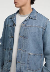 Chaqueta de denim azul claro con cierre de botones en el frente, grandes bolsillos y un cuello clásico. Presenta un acabado desgastado y detalles de costura visibles.