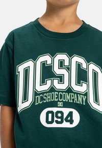 Tmavě zelené tričko s krátkým rukávem s bílým a světle zeleným grafickým textem: "DC Shoe Company" a "094" vepředu. Měkký bavlněný materiál.
