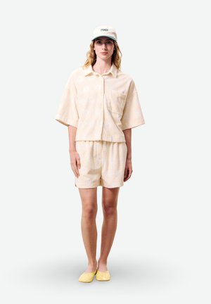 Femme portant une chemise beige à manches courtes à boutons, un short assorti, une casquette blanche avec texte et des chaussures jaunes sans lacets, debout devant un fond blanc.