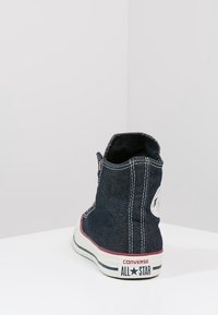 Baskets montantes Converse en denim foncé, avec semelle en caoutchouc blanc, accents rouges et patch circulaire sur le talon extérieur.