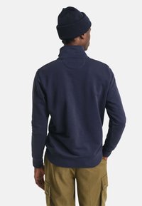 Navyblauwe fleece pullover met een hoge kraag, gestructureerde stof en een cirkelvormige accent aan de achterkant, gecombineerd met khaki cargobroeken.