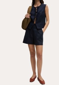 Granatowy lniany bezrękawnik z przodu wiązany, połączony z pasującymi szortami. Brązowa torba na ramię i czerwono-brązowe buty dopełniają stylizację.