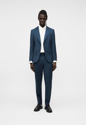 Bărbat în picioare, purtând un costum albastru în carouri, croială strâmtă, cămașă albă și mocasini negri, pe un fundal simplu, deschis la culoare.