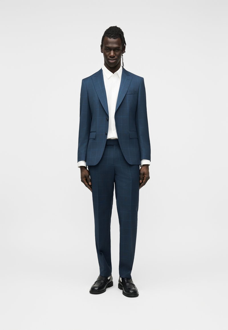 Άνδρας που στέκεται φορώντας ένα εφαρμοστό μπλε καρό κοστούμι με λευκό πουκάμισο και μαύρα μοκασίνια, σε απλό φωτεινό φόντο.