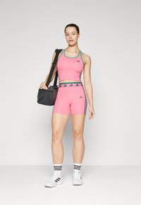 Różowy, sportowy, krótki top i szorty z akcentami w kolorze turkusowym. Czarny logo Adidas na obu elementach. Modelka trzyma czarną torbę sportową, nosi białe buty.