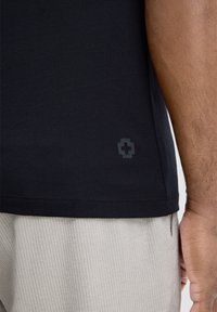 T-shirt en coton noir avec une texture lisse et un petit logo gris au niveau de l'ourlet. Associé à un pantalon à côtes léger.