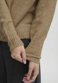 Pull en tricot brun clair avec des poignets côtelés. Texture douce, coupe décontractée et large col. Des mains ajustent l'ourlet tout en portant un pantalon sombre.