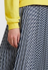 Maglione giallo a coste con maniche lunghe abbinato a una gonna blu navy a pieghe con strisce bianche diagonali. Primo piano di una mano sulla gonna.