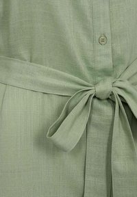 Top de lino verde claro con frente abotonado y cinturón atado en la cintura. La tela tiene un acabado texturizado que realza su diseño casual.