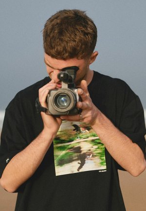 Młody mężczyzna w czarnej koszulce z nadrukiem patrzy przez wizjer kamery wideo, nagrywając na zewnątrz na tle czystego nieba.