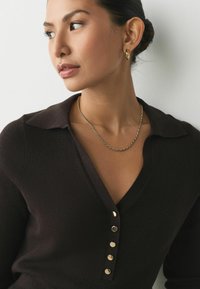 Bruine gebreide top met een kraag, voorzien van vijf metalen knopen en een subtiele ribbelstructuur. Een gouden ketting benadrukt het ontwerp.
