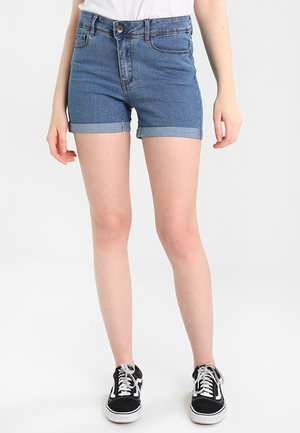 Vrouw die blauwe denimshorts met opgerolde zoom, een wit T-shirt en zwart-witte vetersneakers draagt, staand tegen een effen witte achtergrond.