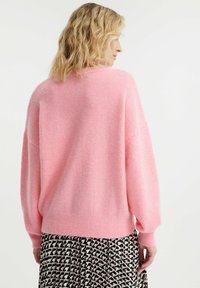 Lichtroze, oversized trui met een zachte, gestructureerde afwerking. Heeft verlaagde schouders en geribbelde manchetten, gecombineerd met een gemusterde rok.
