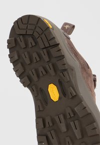 Подметка на обувка с дълбоки грайфери от тъмна гума, жълт лого Vibram в центъра, кафяв велурен горен слой с видима дръжка за издърпване.