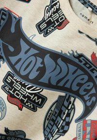 Camiseta de algodón beige con logotipos y gráficos coloridos y variados, incluyendo "Hot Wheels". El diseño incluye acentos en azul y rojo.