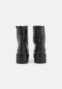 Paire de bottines en cuir noir à talons carrés, vues de dos sur un fond blanc.