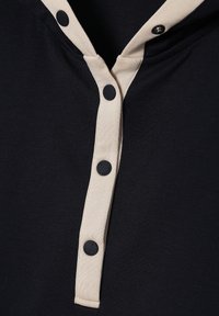 Gros plan d'une chemise en tissu noire avec une bande de boutons beige et des boutons-pression noirs, dont trois sont fixés.