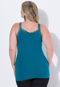Donna con lunghi capelli biondi che indossa un top senza maniche di pizzo turchese con bordo, pantaloni neri e orecchini neri pendenti, girata di spalle su uno sfondo bianco.