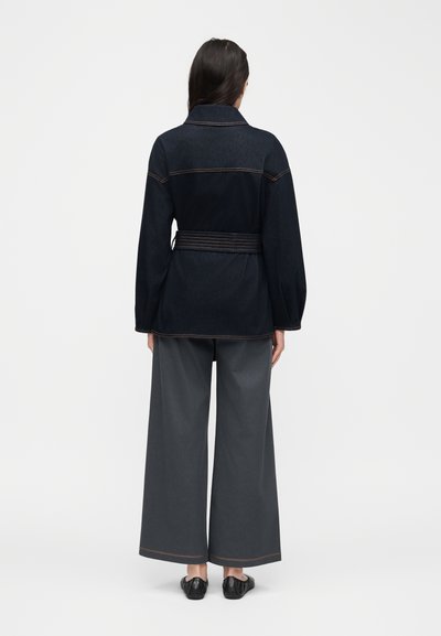 Γυναίκα που στέκεται πλάτη φορώντας μαύρο σακάκι με ζώνη, παντελόνι φαρδύ γκρι σκούρο και μαύρα επίπεδα παπούτσια σε απλό φόντο.