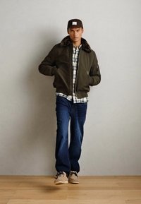 Oliven grøn bomberjakke med pelskrave, lagd over en ternet skjorte, parret med blå jeans og beige sneakers. Indstillingen er minimalistisk.