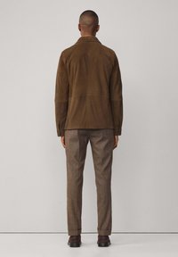 Braune Wildlederjacke mit einem klassischen Kragen und einem Rückenteil mit Steppdetails, kombiniert mit gemusterten braunen Hosen und dunklen Schuhen.