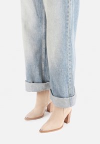 Botas de tobillo de ante beige con puntera afilada, detalle en plata y tacón de bloque de madera, combinadas con vaqueros de denim azul claro arremangados.