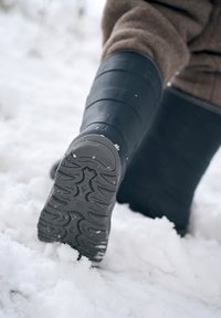Botte en caoutchouc noire avec semelle texturée, présentant des crans et des motifs pour l'adhérence, partiellement immergée dans la neige. L'arrière-plan montre de la neige floue.