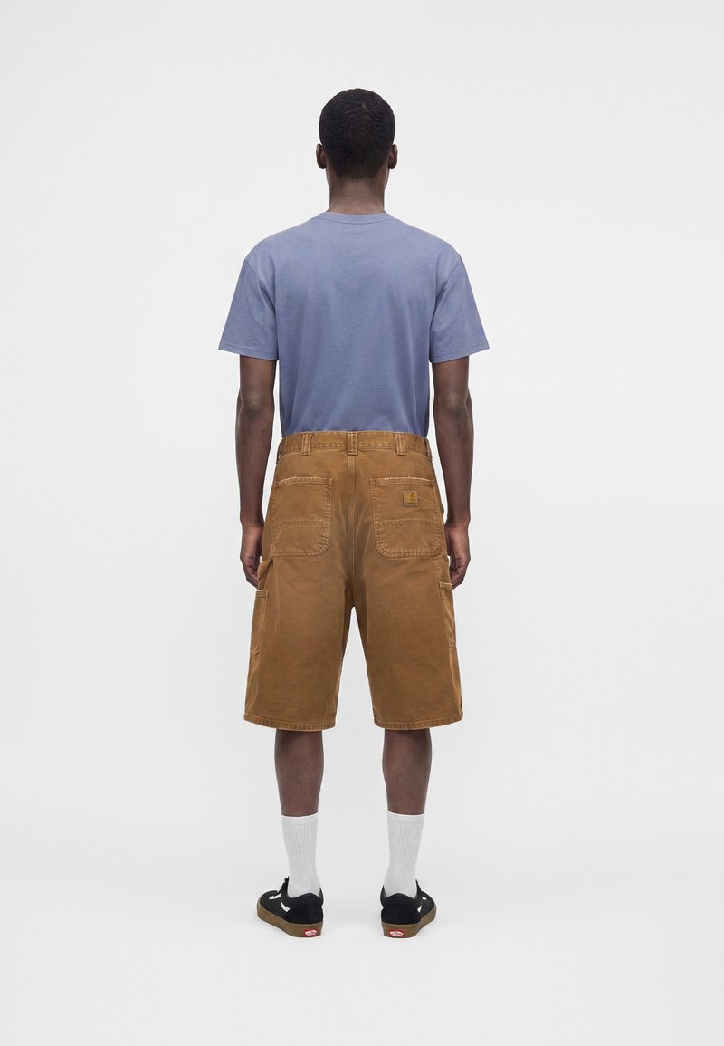 Hombre con camiseta azul, pantalones cortos marrones hasta la rodilla con múltiples bolsillos, calcetines blancos y zapatillas negras, de pie de espaldas sobre fondo blanco.