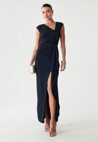Navy wieczorowa sukienka z rękawami typu cap, marszczonym korpusem i dekoltem do uda. Gładki, błyszczący materiał doskonale komponuje się z srebrnymi sandałami na obcasie.