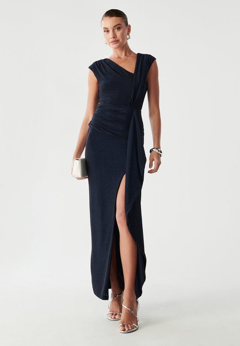 Navy wieczorowa sukienka z rękawami typu cap, marszczonym korpusem i dekoltem do uda. Gładki, błyszczący materiał doskonale komponuje się z srebrnymi sandałami na obcasie.
