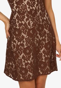 Robe en dentelle brune avec motifs floraux, tissu transparent, et un design ajusté avec une silhouette évasée. Les détails incluent des coutures complexes.