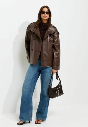 Vrouw draagt een oversized bruine leren jas, een donkere trui, blauwe wijde jeans, zwarte sandalen, zonnebril, houdt een kleine bruine handtas vast.