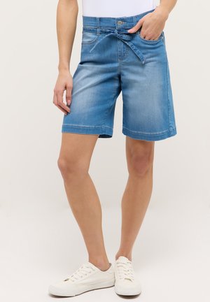 Mujer con shorts de mezclilla azul de longitud media con cinturón anudado en la cintura y zapatillas blancas, de pie frente a un fondo liso.